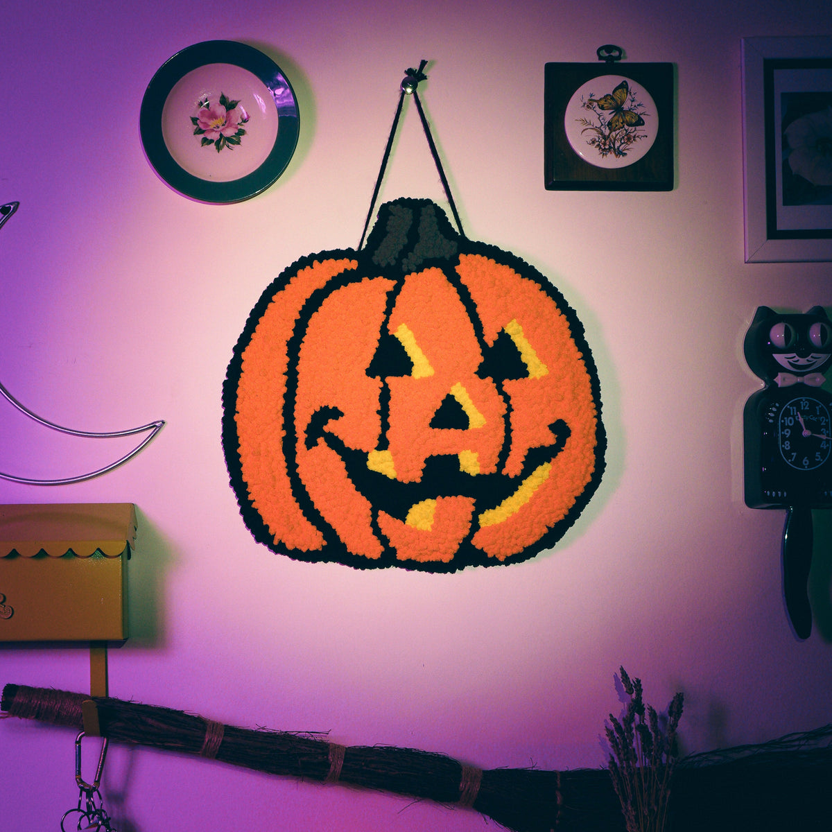 a jack-o'-lantern punch needle wall hanging hanging on a gallery wall with witchy items arranged around it.