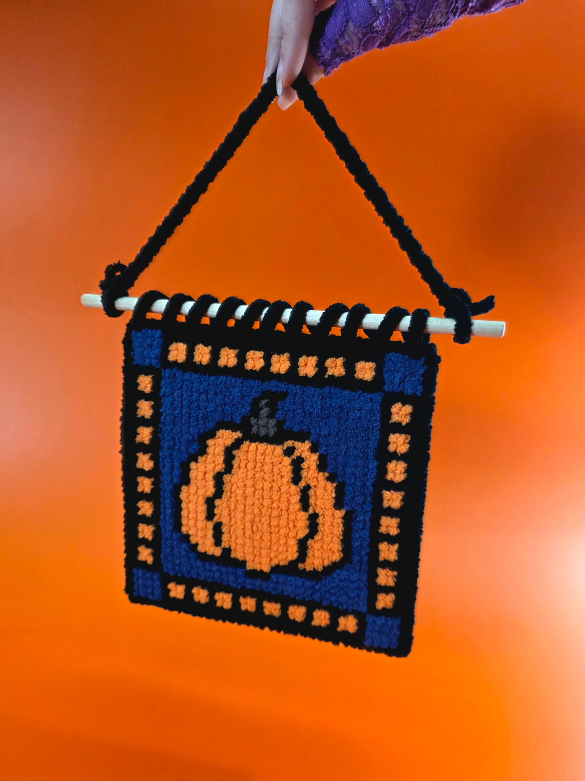Hand holding up a pumpkin needlepoint wall hanging with a blue background and orange and black squares around the edge.