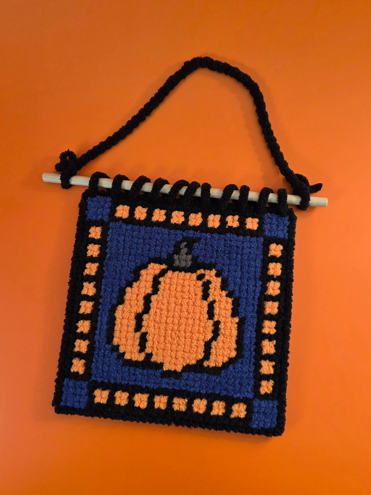 pumpkin needlepoint wall hanging with a blue background and orange and black squares around the edge.