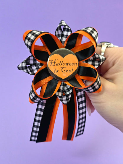 Prize ribbon made with blac, orange, and checkered black and white ribbon has an orange button in the center that says "Halloween is cool". A hand is holding it in front of a purple background.