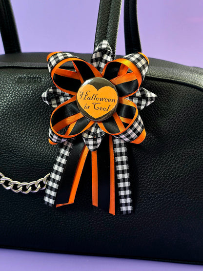 Prize ribbon made with blac, orange, and checkered black and white ribbon has an orange button in the center that says "Halloween is cool". It is attached to a bag.