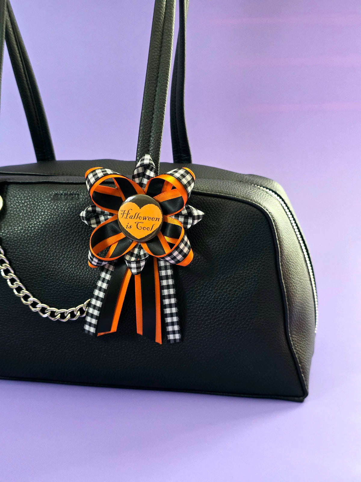 Prize ribbon made with blac, orange, and checkered black and white ribbon has an orange button in the center that says "Halloween is cool". It's attached to a bag.