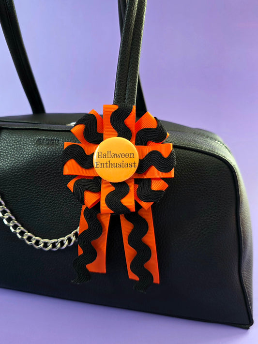A prize ribbon rosette made with orange and black ribbon with an orange button in the center that says "Halloween Enthusiast". It is attached to a bag.