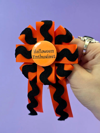 A prize ribbon rosette made with orange and black ribbon with an orange button in the center that says "Halloween Enthusiast". It is being held up by a hand in front of a purple backdrop.