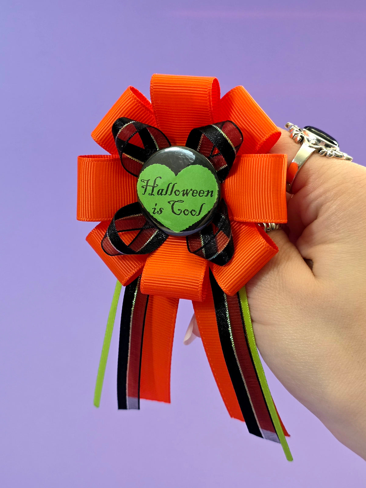 a prize ribbon rosette made of orange, green, and black ribbon with a green button in the center that says "Halloween is cool" with a black heart around it. A hand it holding it up in front of a purple backdrop.
