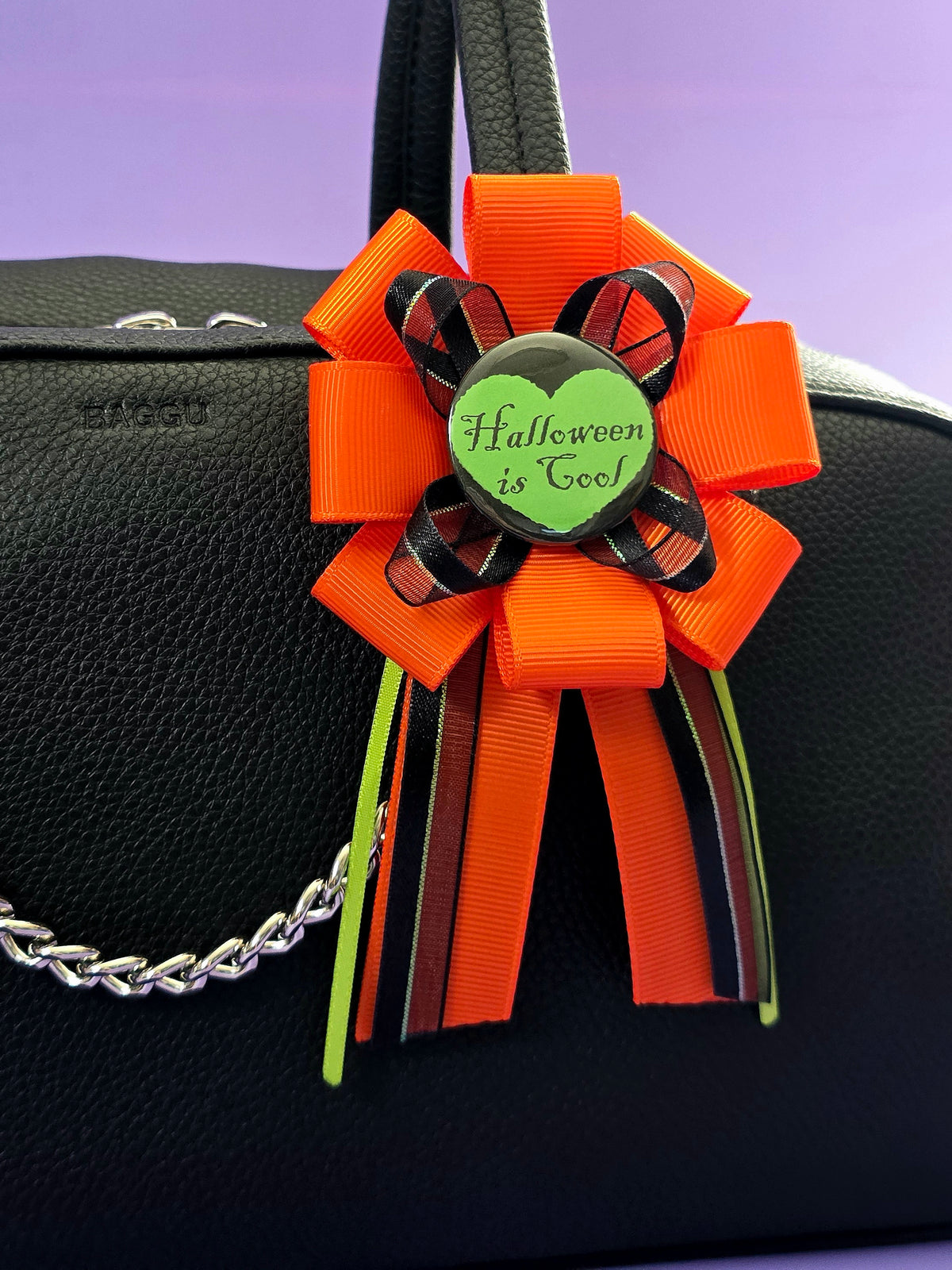 a prize ribbon rosette made of orange, green, and black ribbon with a green button in the center that says "Halloween is cool" with a black heart around it. it is attached to a purse here.