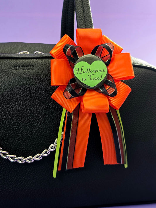 a prize ribbon rosette made of orange, green, and black ribbon with a green button in the center that says "Halloween is cool" with a black heart around it. it is attached to a purse here.
