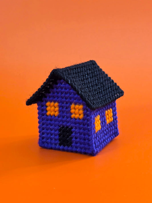 a three dimensional small haunted house tchotchke  made from plastic canvas and needlepoint. The walls are made from purple yarn, the windows from orange yarn, and the roof and door from black yarn.