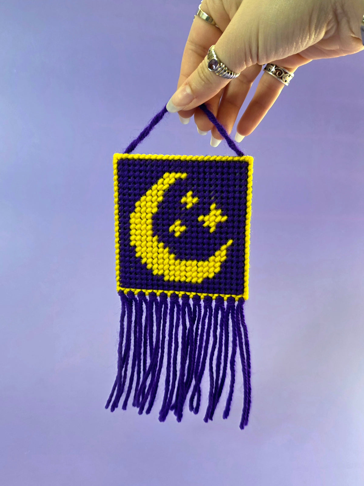 a needlepoint wall hanging with a yellow moon and stars with a purple background being held up in front of a purple background