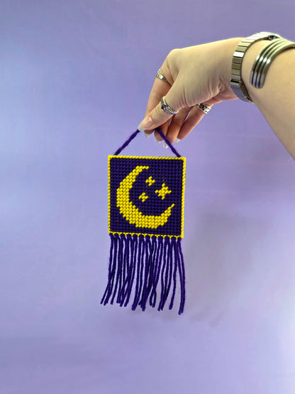a needlepoint wall hanging with a yellow moon and stars with a purple background being held up in front of a purple background