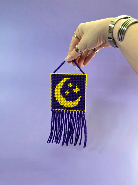 a needlepoint wall hanging with a yellow moon and stars with a purple background being held up in front of a purple background