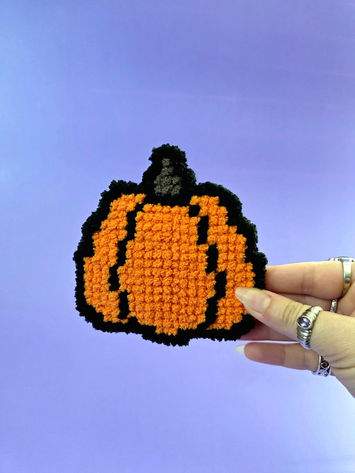 Small fluffy needlepoint pumpkin being held in front of a purple backdrop