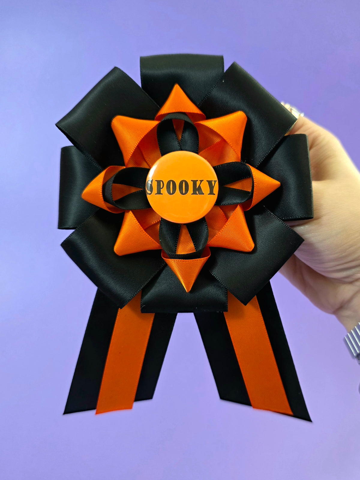 Prize ribbon rosette made with orange and black ribbons, it has an orange button in the center that says "spooky". It is being held by a hand in front of a purple backdrop.