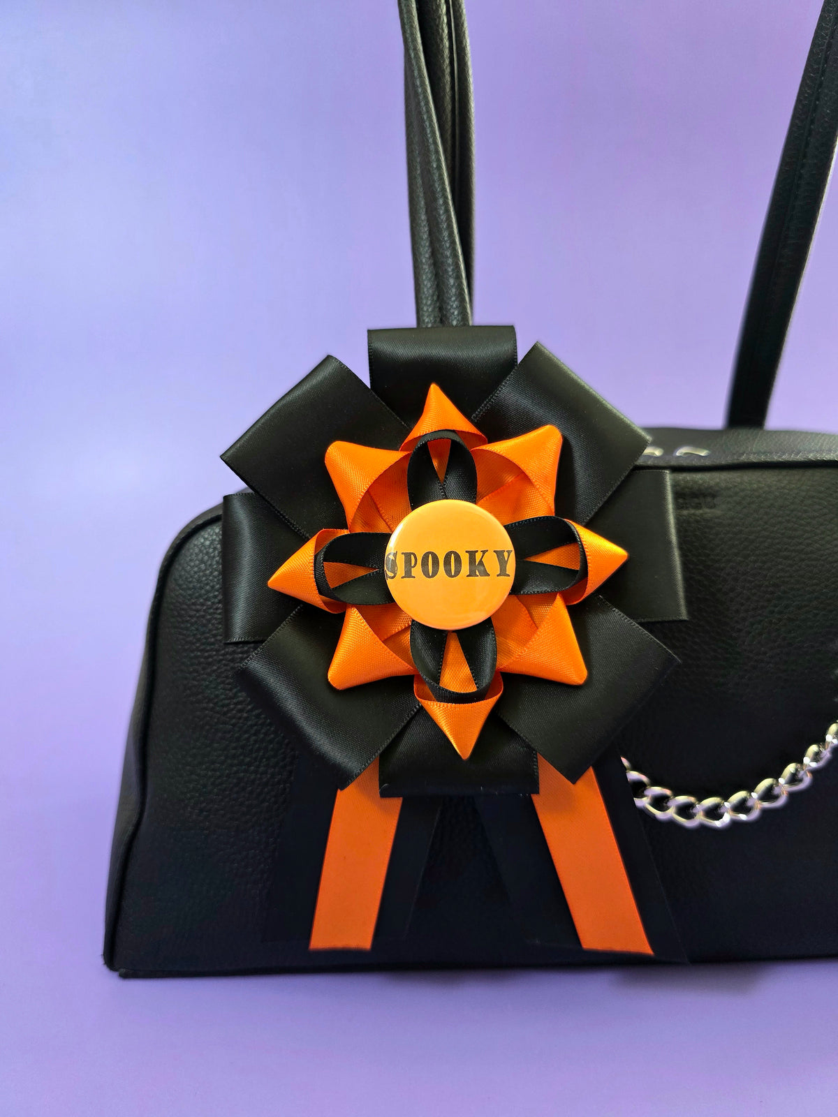 Prize ribbon rosette made with orange and black ribbons, it has an orange button in the center that says "spooky". It is attached to a bag.