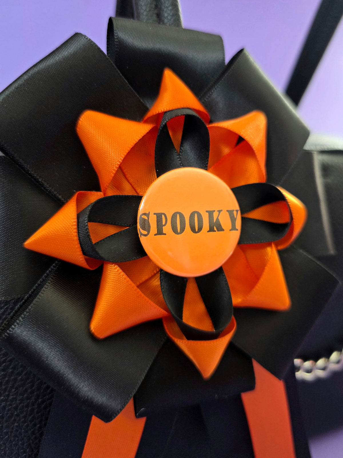 Prize ribbon rosette made with orange and black ribbons, it has an orange button in the center that says "spooky". Close up photo.